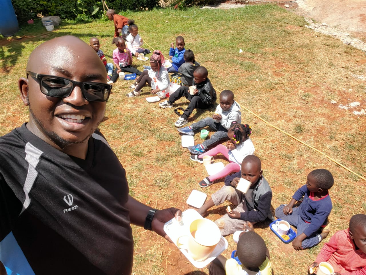 Local leader Joe with smiling children enjoying a meal under the bright Kenyan sun, part of RDM.life's community nourishment program.